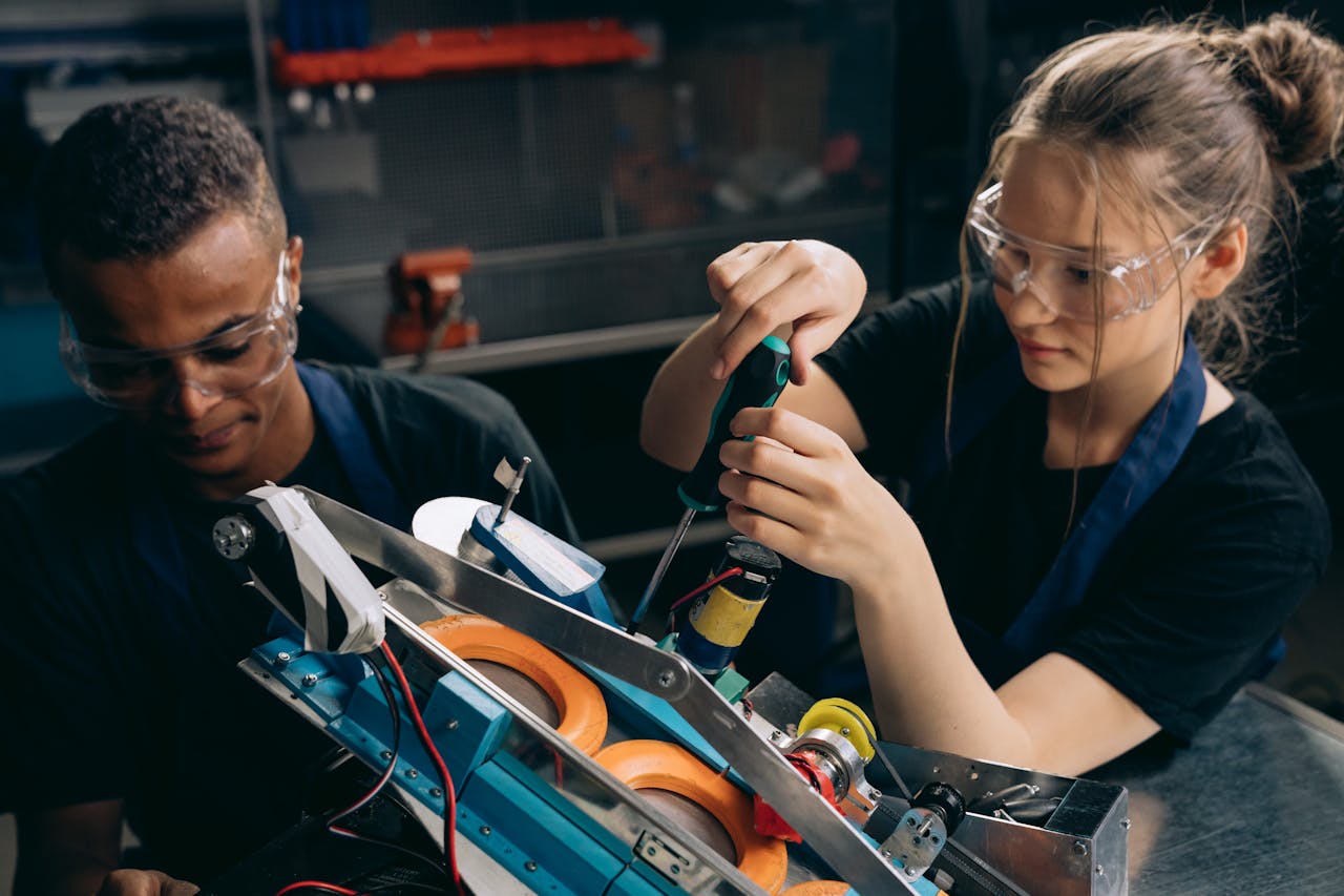 our-mission Two students working together on building a robot in an industrial setting, wearing protective goggles.